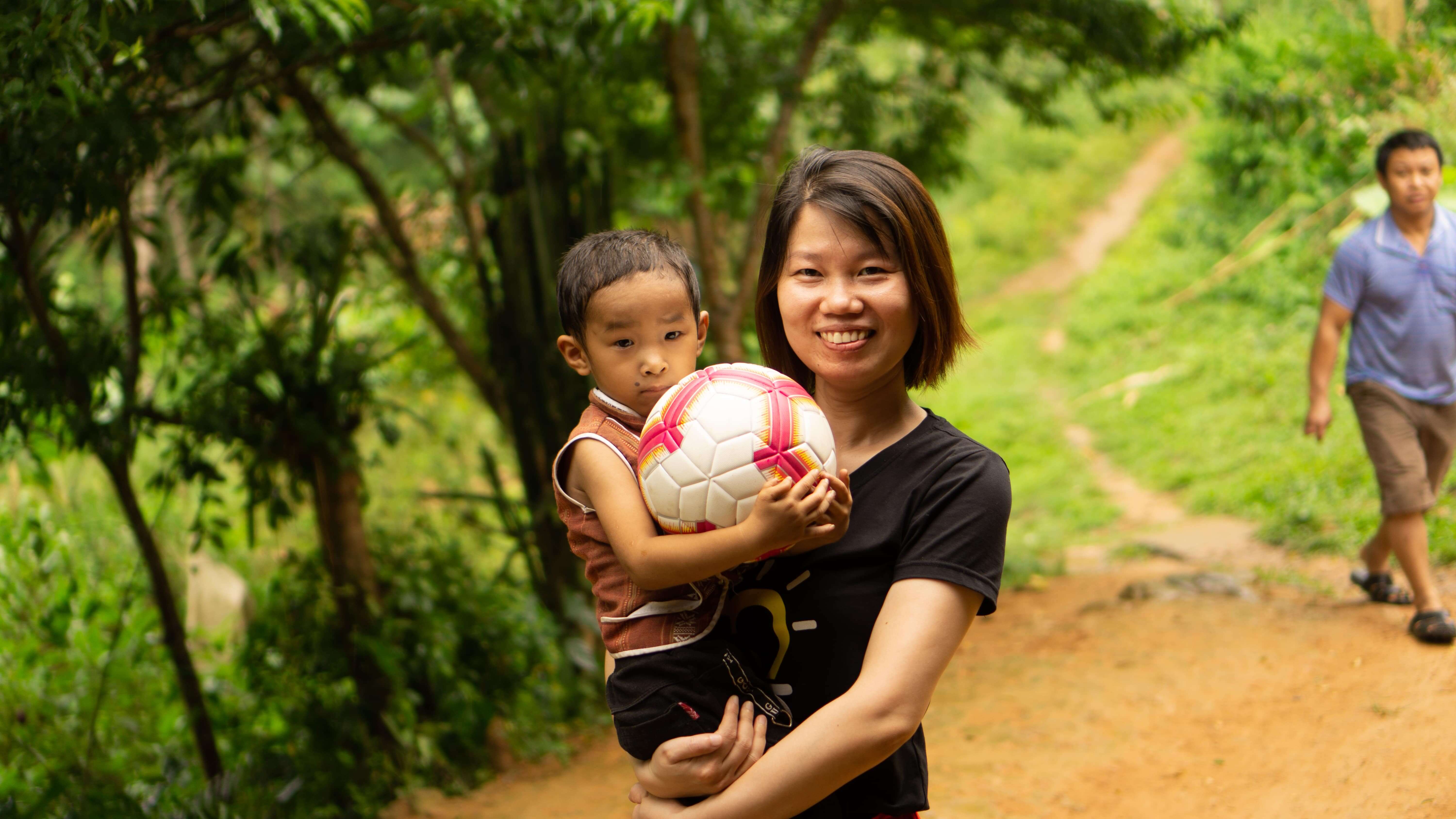 Woman smiling holding child with soccer ball outdoors on dirt path