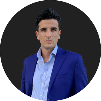 A man wearing a blue shirt and a suit stands against a dark background.