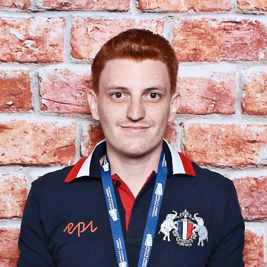Red-haired man wearing navy polo with crest and event lanyard against brick wall